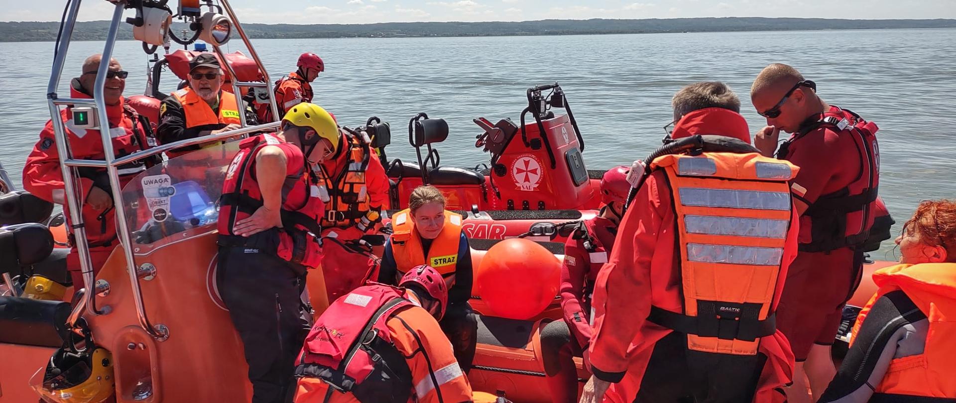Na wodzie ustawione dwie łodzie ratownicze w nich ratownicy w czerwonych kombinezonach. W oddali, na horyzoncie brzeg. Niebieskie niebo.