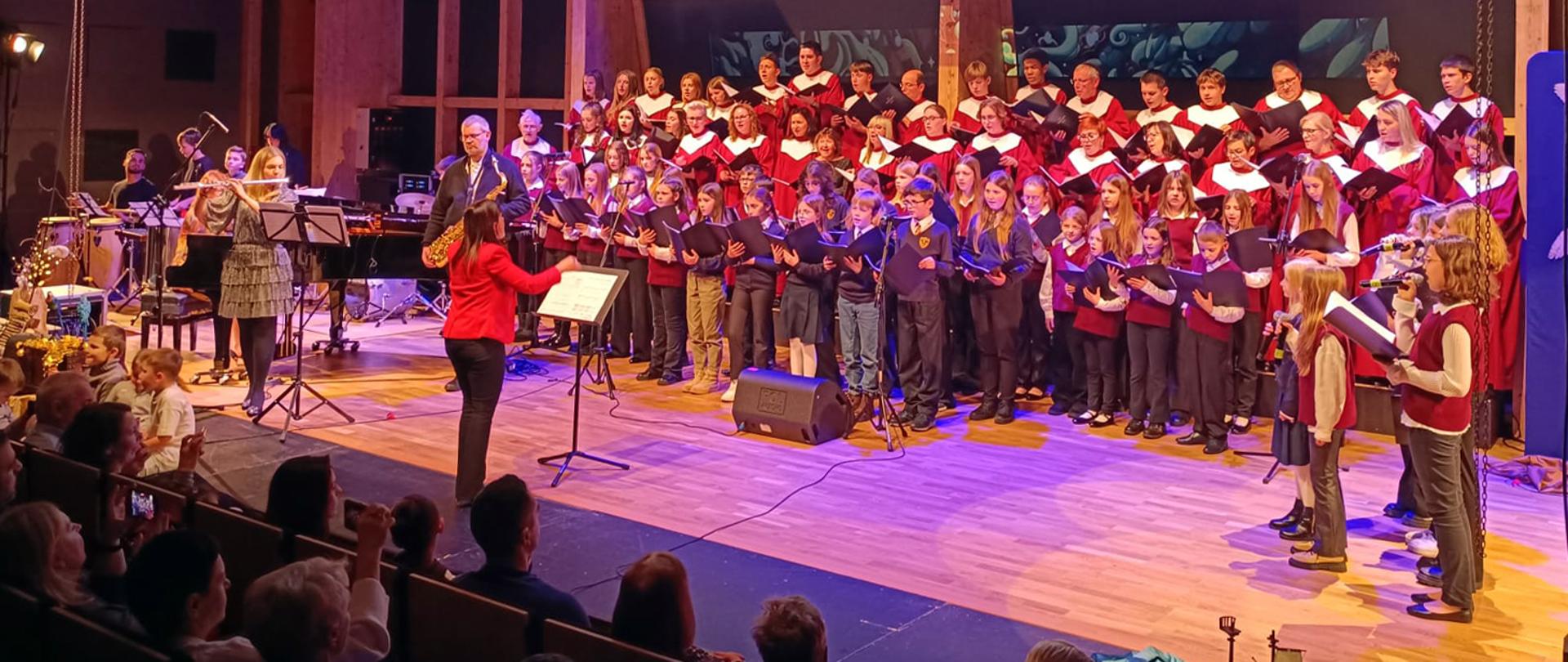 Duża grupa osób stoi na scenie, w rękach trzymają nuty, przed nimi stoi dyrygentka, z lewej strony widać osoby grające na różnych instrumentach muzycznych, w lewym dolnym rogu zdjęcia widać osoby z widowni.