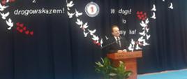 Wiceminister Włodzimierz Bernacki stoi przy mównicy. Przy mównicy są donice z kwiatami. W tle grantowa zasłona. Na niej wycięte z papieru ptaki i serca. Widoczny fragment większego napisu. 