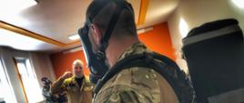 Policjant ubrany w aparat ochrony dróg oddechowych przed nim stoi strażak który omawia sprzęt obok na stołach leży sprzęt dalej ubierani są inni policjanci. 