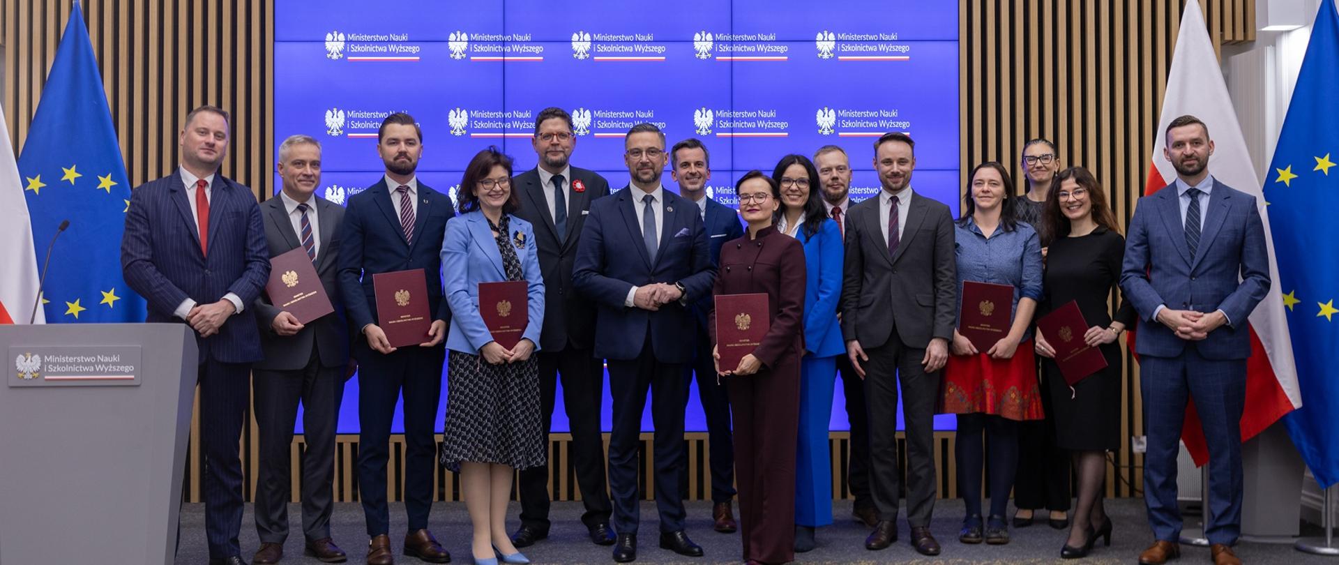 Pod ścianą z cienkich pionowych drewnianych listewek stoi grupa kilkunastu osób, niektórzy trzymają ciemnoczerwone teczki, pośrodku grupy minister Kulasek i wiceminister Szeptycki, za nimi wbudowany w ścianę wielki niebieski ekran, po bokach pod ścianą flagi Polski i UE.