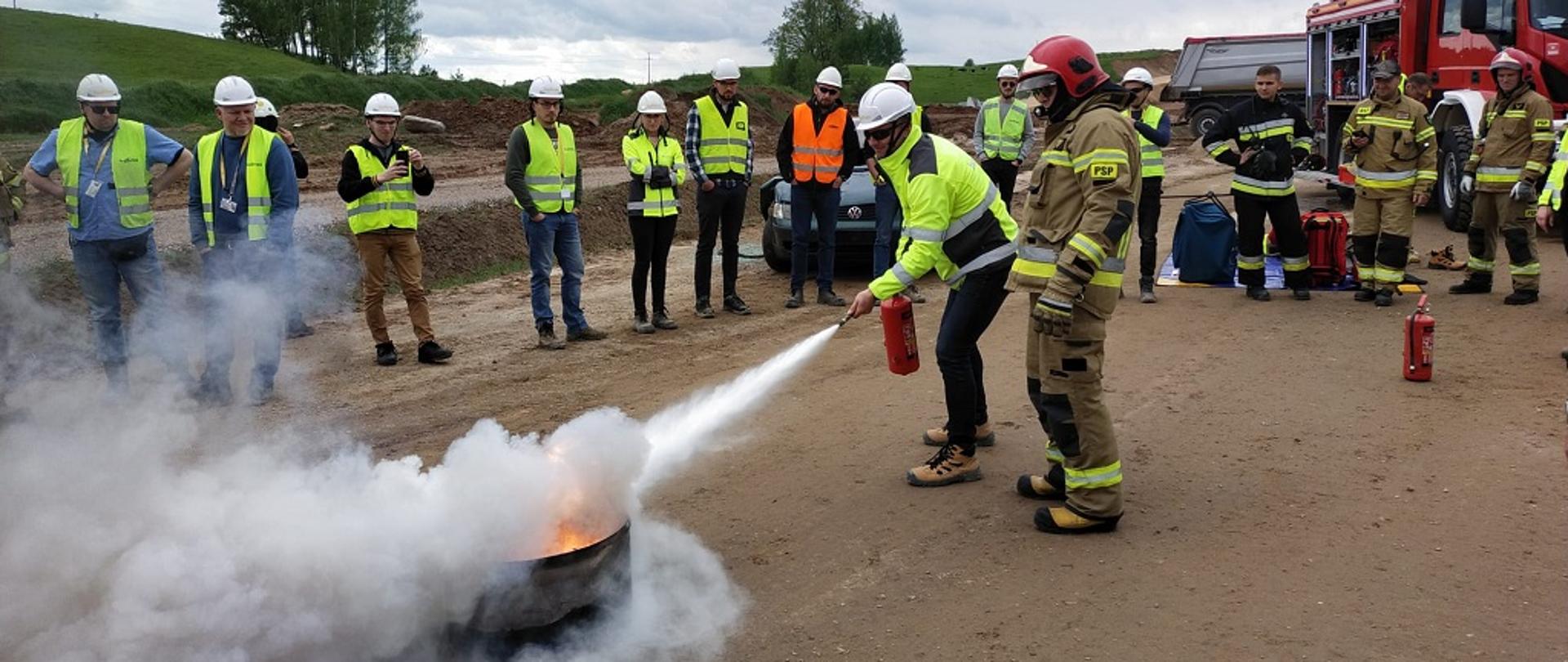 Pracownicy budowlani w asyście strażaka gaszą ogień w beczce. 
