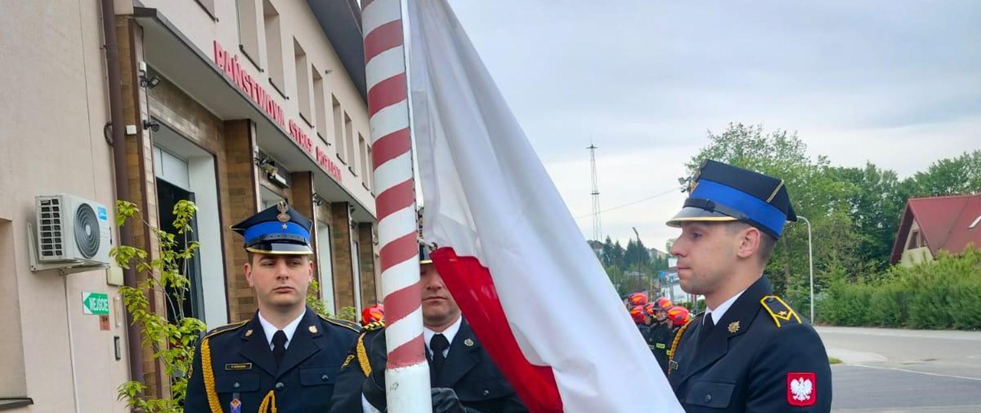 Zdjęcie przedstawia strażaków KP PSP Lesko podczas uroczystego wciągnięcia na maszt flagi Rzeczpospolitej Polskiej.
