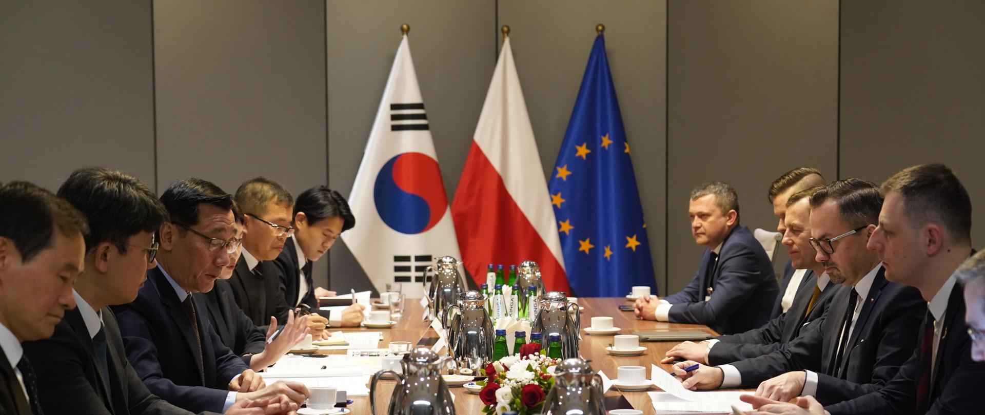 Grupa osób siedzi po obu stronach długiego stołu. Z tyłu stoją flagi Korei Południowej, Polski i Unii Europejskiej.