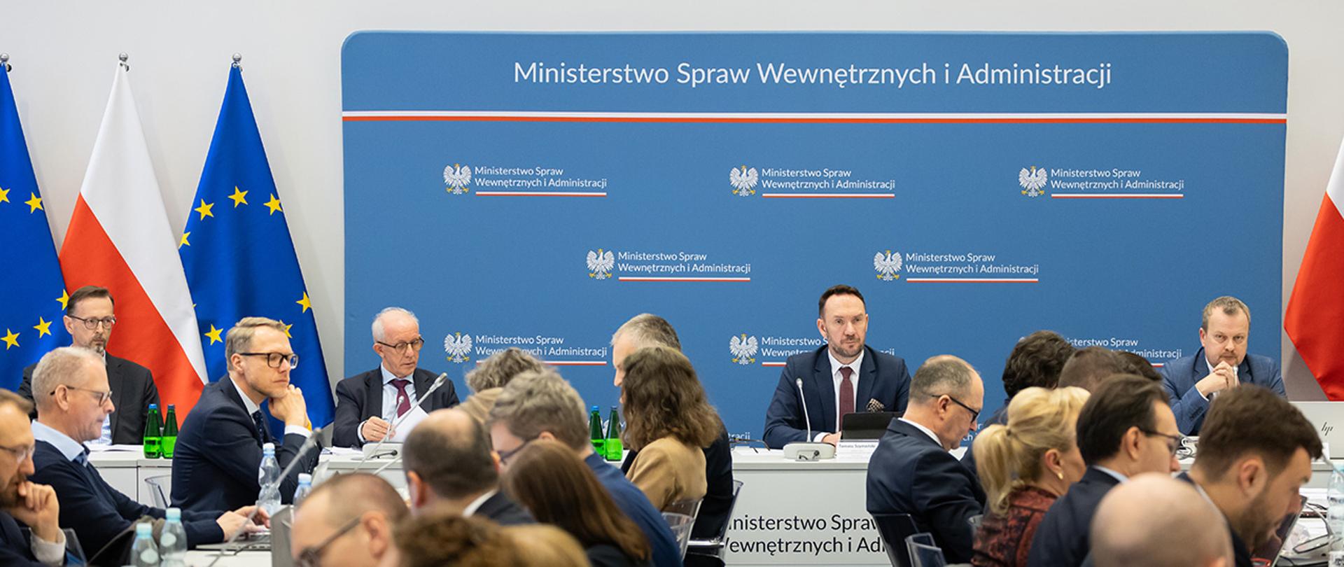 Oficjalne spotkanie w MSWiA. Wiceminister Tomasz Szymański zasiada przy stole prezydialnym na tle logo ministerstwa. Wokół niego uczestnicy zebrania, część przegląda dokumenty, inni słuchają.