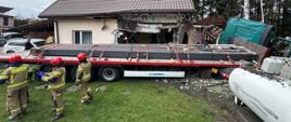 Na zdjęciu widoczna jest scena po wypadku drogowym z udziałem ciężarówki, która uderzyła w budynek mieszkalny. Do zdarzenia doszło na terenie posesji, na co wskazują zniszczony fragment ściany domu oraz obecność trawnika i drzew.
Ciężarówka z naczepą, na której przewożone są długie, ciemne elementy (prawdopodobnie metalowe profile), częściowo znajduje się na terenie posesji, a jej kabina jest przechylona w stronę uszkodzonego budynku. Ściana domu, w którą uderzyła ciężarówka, jest poważnie zniszczona, widać dziurę i gruz.
Wokół miejsca zdarzenia zgromadziła się grupa strażaków w czerwonych hełmach i beżowych mundurach z żółtymi pasami. Niektórzy z nich stoją w pobliżu ciężarówki, inni przyglądają się uszkodzeniom budynku. Na trawniku leżą fragmenty zniszczonej ściany i inne elementy.
W tle widoczny jest inny budynek, drzewa oraz pochmurne niebo. Całość sceny sugeruje poważny wypadek, który spowodował znaczne uszkodzenia zarówno pojazdu, jak i budynku, i wymaga interwencji służb ratowniczych.