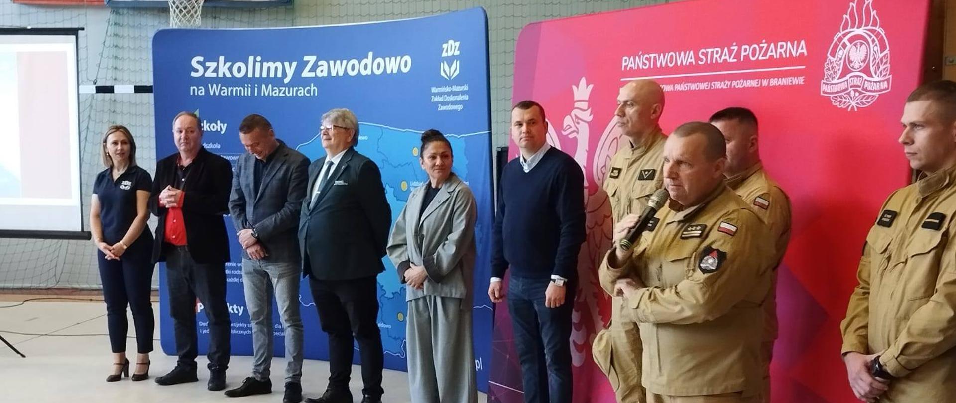 Zdjęcie przedstawia oficjalne wydarzenie z udziałem przedstawicieli Państwowej Straży Pożarnej oraz Warmińsko-Mazurskiego Zakładu Doskonalenia Zawodowego (ZDZ). Po prawej stronie stoją funkcjonariusze PSP w umundurowaniu specjalnym, na tle czerwonej ścianki z logiem i nazwą Komenda Powiatowa PSP w Braniewie. Jeden z nich (z mikrofonem) prawdopodobnie przemawia, pełniąc funkcję prowadzącego. Po lewej stronie stoją cywilni przedstawiciele – kobieta w koszulce z logo ZDZ oraz kilku mężczyzn i kobiet, którzy wyglądają na reprezentantów instytucji współpracujących, władz lokalnych lub edukacyjnych.