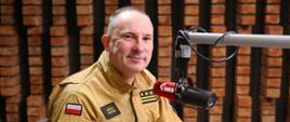 Strażak Państwowej Straży Pożarnej w mundurze służbowym siedzi przy stole w studiu radiowym. Przed nim znajduje się mikrofon z logo Radia Kielce oraz dokumenty, które podpisuje długopisem. Mężczyzna uśmiecha się do aparatu, a w tle widoczna jest ściana z cegły i elementy wyposażenia studia nagraniowego.