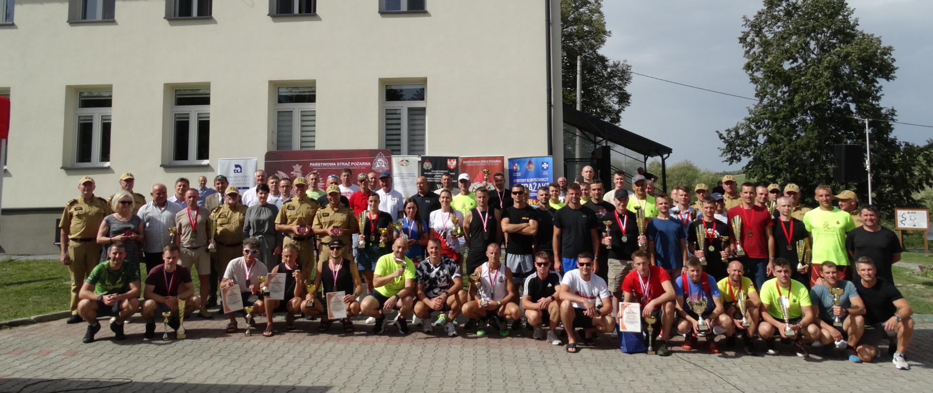Duża grupa strażaków oraz uczestników biegu górskiego stoi przed budynkiem komendy PSP. Na pierwszym planie widoczni są zawodnicy z medalami, dyplomami i pucharami, a w tle banery organizatorów wydarzenia.