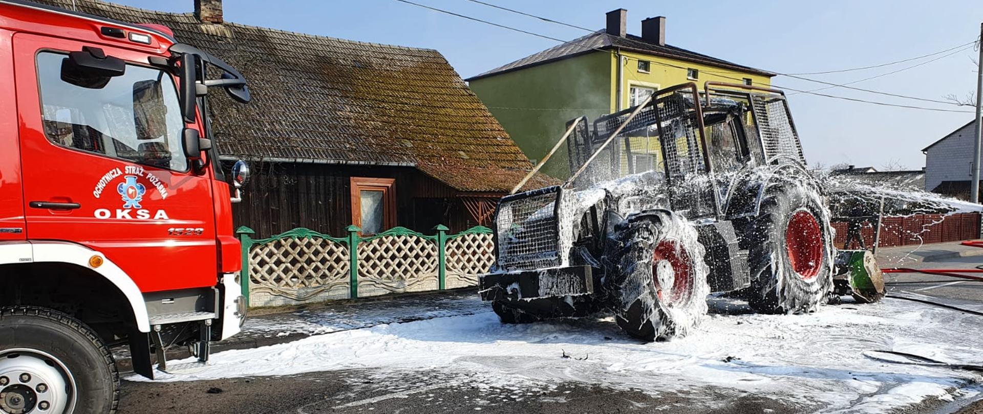Zdjęcie po lewej stronie przedstawia przód samochodu ratowniczo-gaśniczego. Na jego drzwiach widniej napis „OSP Oksa”. W środkowej części zdjęcia widać spalony ciągnik rolniczy pokryty warstwą piany gaśniczej. Zarówno pojazd strażacki jak i ciągnik znajdują się na pasie jezdni drogi wojewódzkiej. W tle widać parterowy, drewniany budynek mieszkalny.
