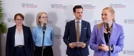 Na tle białej ścianki z napisami Ministerstwo Edukacji Narodowej stoi trzy kobiety i mężczyzna. Przed nimi ustawione na statywach dwa mikrofony, przed jednym z nich stoi minister Nowacka i mówi. 