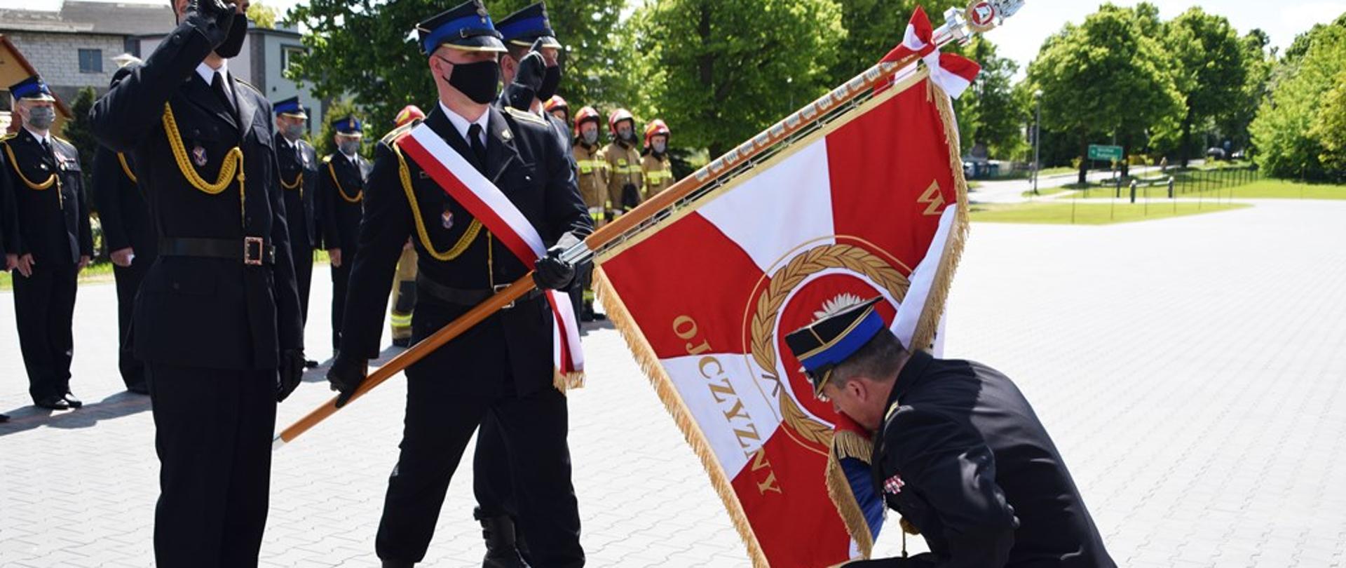 Na zdjęciu komendant powiatowy klęka na kolano przed sztandarem i całuje jego płat. Żegna się, odchodząc ze służby. Przed nim poczet sztandarowy, w tle załoga. Wszyscy umundurowani galowo. Uroczystość na placu przed budynkiem.