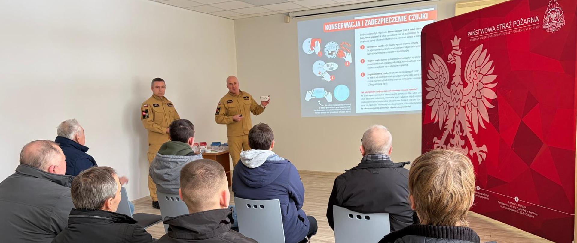 Strażacy stoją i omawiają mieszkańcom miasta zasady instalowania czujek dymu i tlenku węgla. Strażak w ręku trzyma czujki. 