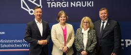 Pod granatowym banerem z napisem Europejskie Targi Nauki stoją cztery osoby, dwóch mężczyzn w szarych garniturach, wiceminister Zioło-Pużuk i kobieta w ubraniu w biało-czarny wzorek.