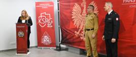 W sali uroczystej stoi kobieta przy mównicy, wygłaszając przemówienie. Obok niej ustawione są dwa czerwone banery Państwowej Straży Pożarnej i KSRG. Po prawej stronie stoją dwaj strażacy: jeden w mundurze piaskowym, drugi w mundurze galowym. W tle widnieje duża czerwona grafika z białym orłem.
