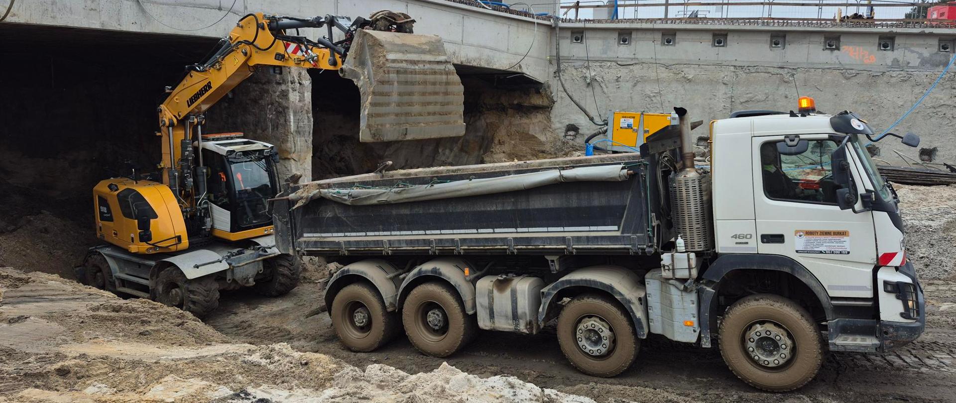 Wywożenie ziemi z tunelu, na pierwszym planie koparka ładuje ziemię na ciężarówkę przy wjeździe do tunelu