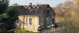 Zdjęcie przedstawia dom jednorodzinny z uszkodzonym dachem i strażaków na drabinie. Nie widać dymu ani ognia.