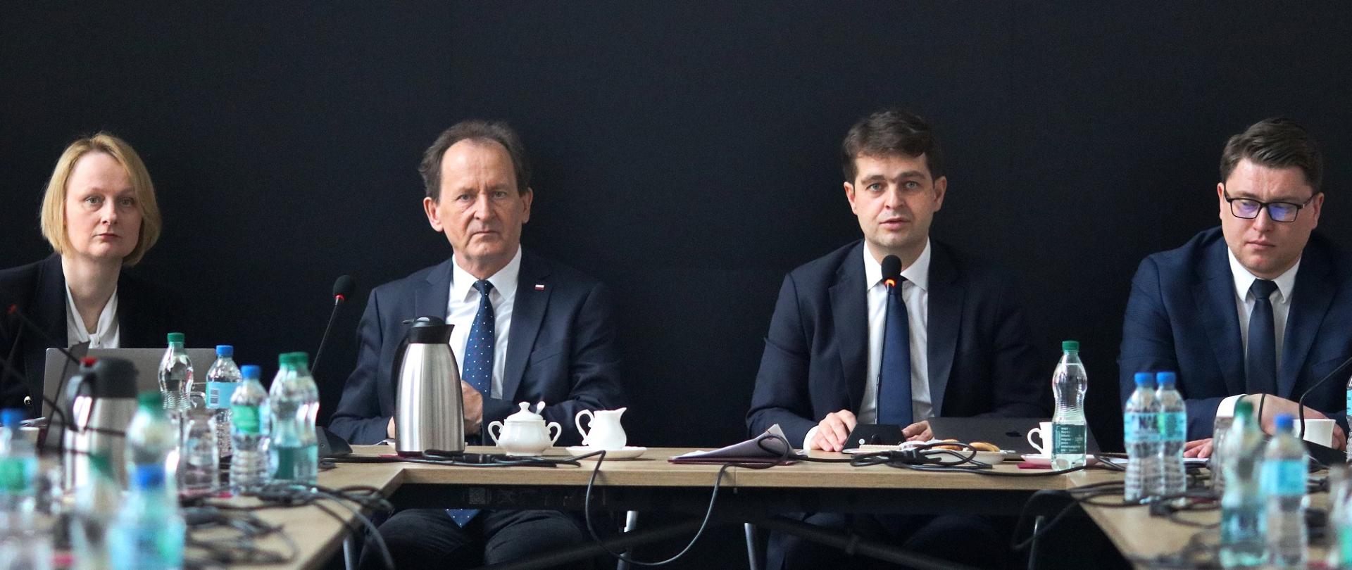 Widok na szczyt stołu stojącego w eleganckiej sali, siedzi trzech mężczyzn w garniturach i kobieta w czarnej marynarce, za nimi czarna ściana.