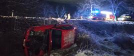 Zdjęcie przedstawia miejsce wypadku czerwonego samochodu osobowego w porze nocnej. Samochód leży na lewym boku, poza drogą. Na drodze stoją samochody: straży pożarnej, policji oraz karetki z łączonymi światłami na drodze stoją funkcjonariusze.