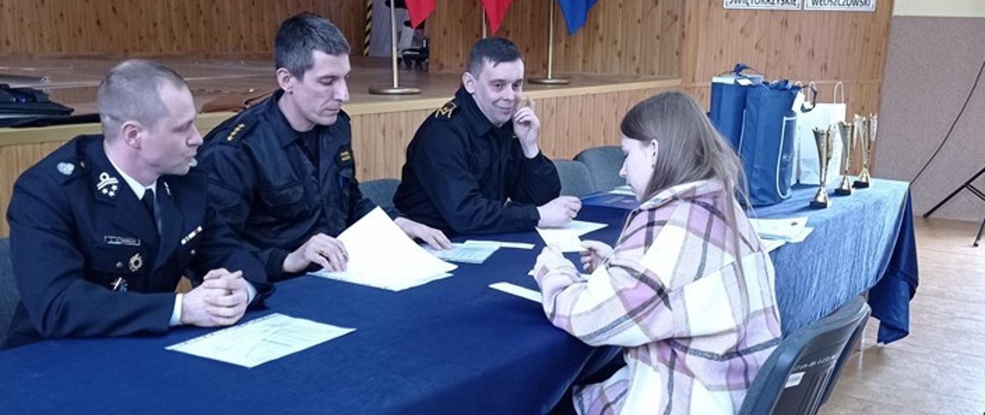 Etap ustny konkursu, sala narad w starostwie powiatowym. Trzyosobowa komisja konkursowa siedzi przy stole prezydialnym, przed nimi uczestniczka konkursu odpowiada na pytania z wylosowanego zestawu.