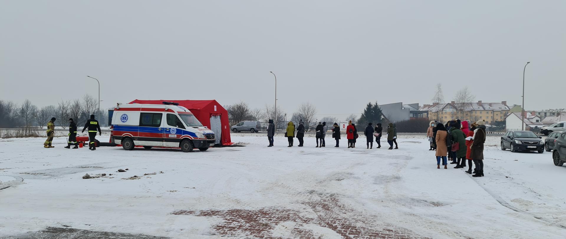 Kolejka ludzi na namiotu strażackiego. Ambulans obok namiotu. Strażacy w tle.