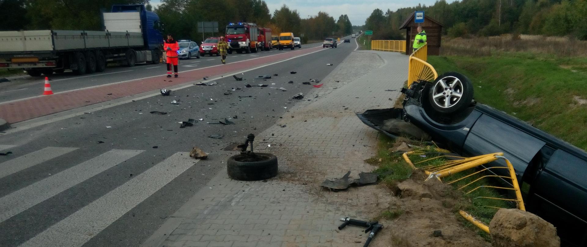 Zdjęcie przedstawia miejsce wypadku - pop prawej wywrócony na dach opel Signum lezy w rowie melioracyjnym. Wokół samochodu są porozrzucane części karoserii, oraz powyginana barierka ochronna. W oddali widać zablokowaną jezdnię oraz służby ratownicze.