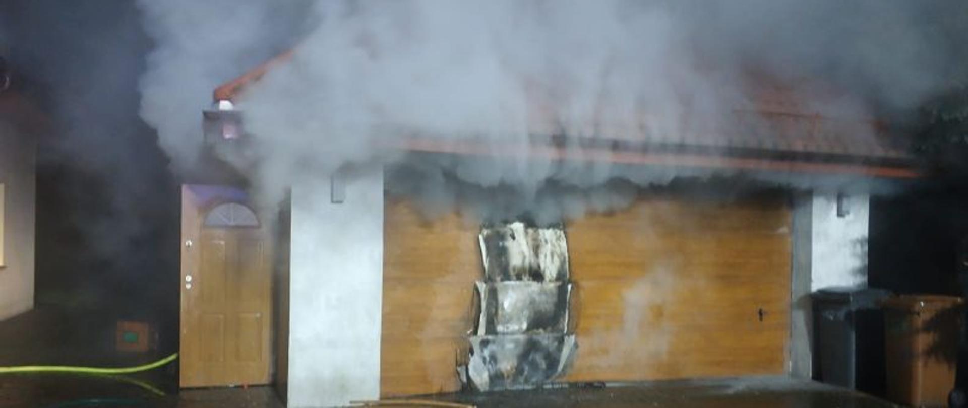 Zdjęcie wykonane w nocy. Centralny punkt stanowi garaż i nadpalona i wygięta od temperatury brama garażowa. Z garażu wydostaje się dym. Przed garażem stoją sanki. Wewnątrz garażu panuje pożar, który powoli zaczyna wydostawać się na zewnątrz budynku. Z lewej strony otwarte drzwi do garażu a z prawej kosze na śmieci