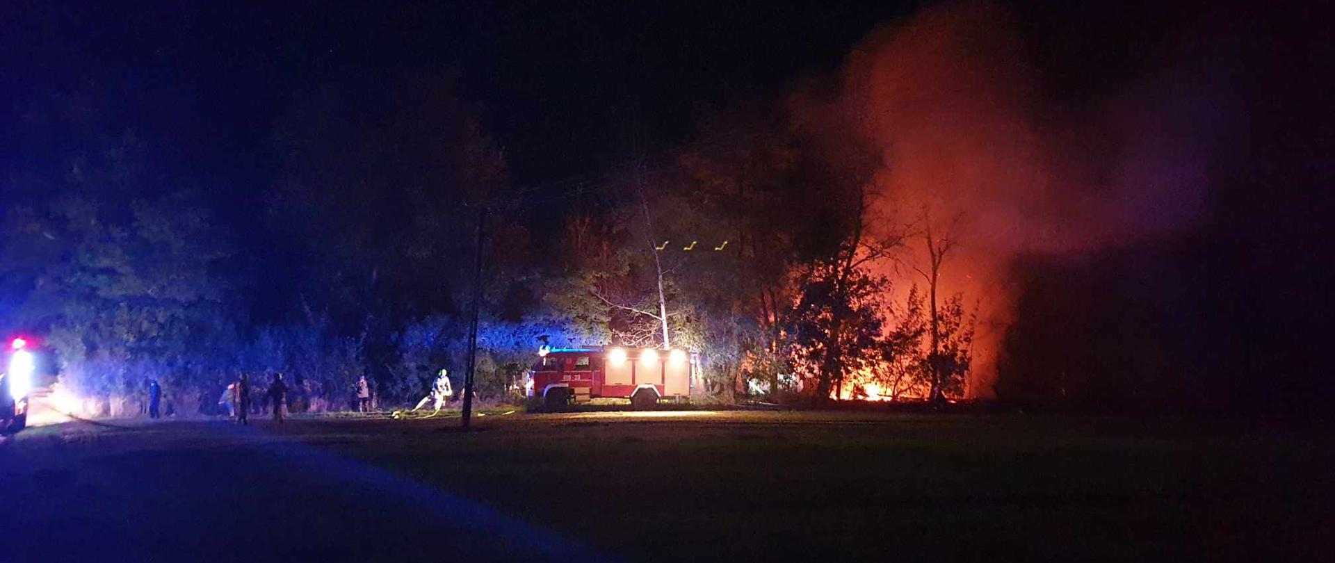  Na zdjęciu widoczna łuna ognia za drzewami oraz samochód ratowniczo-gaśniczy. Koło wozu strażackiego zarysowane sylwetki strażaków rozwijających węże pożarnicze. Zdjęcie zrobione w porze nocnej.