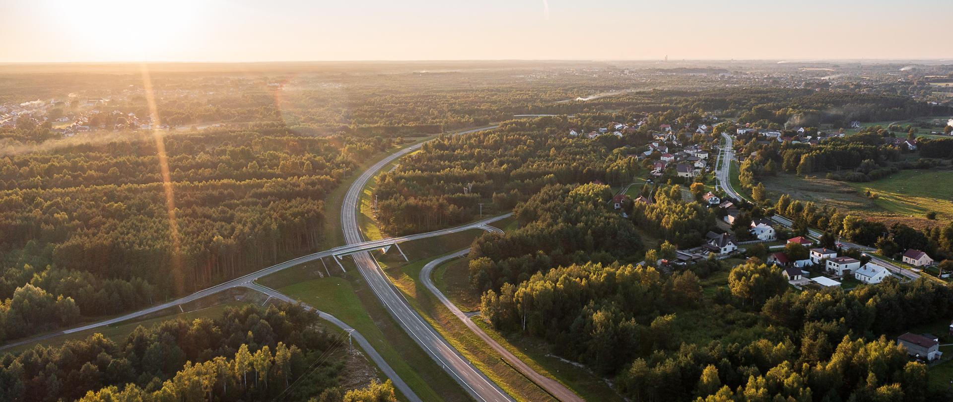 Popołudniowa pora dnia, świeci słońce. Widok z lotu ptaka na obwodnicę Stalowej Woli i Niska. Droga położona w terenie leśnym, skręca delikatnie po łuku w prawo. Nad drogą zlokalizowany jest wiadukt drogowy. Po prawej stronie, za drzewami widać budynki. 