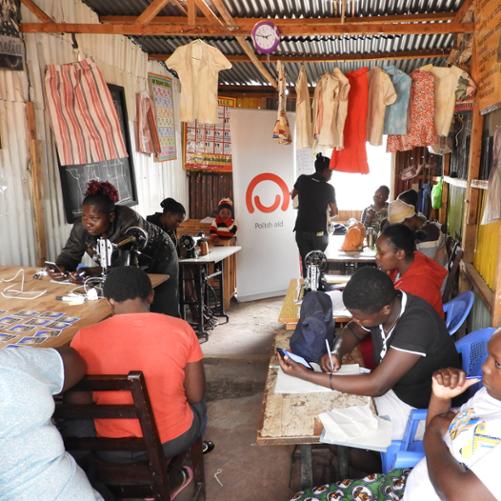 grupa Kenijek w małym pomieszczeniu z maszynami do szycia. na ścianach pomieszczenia rysunki