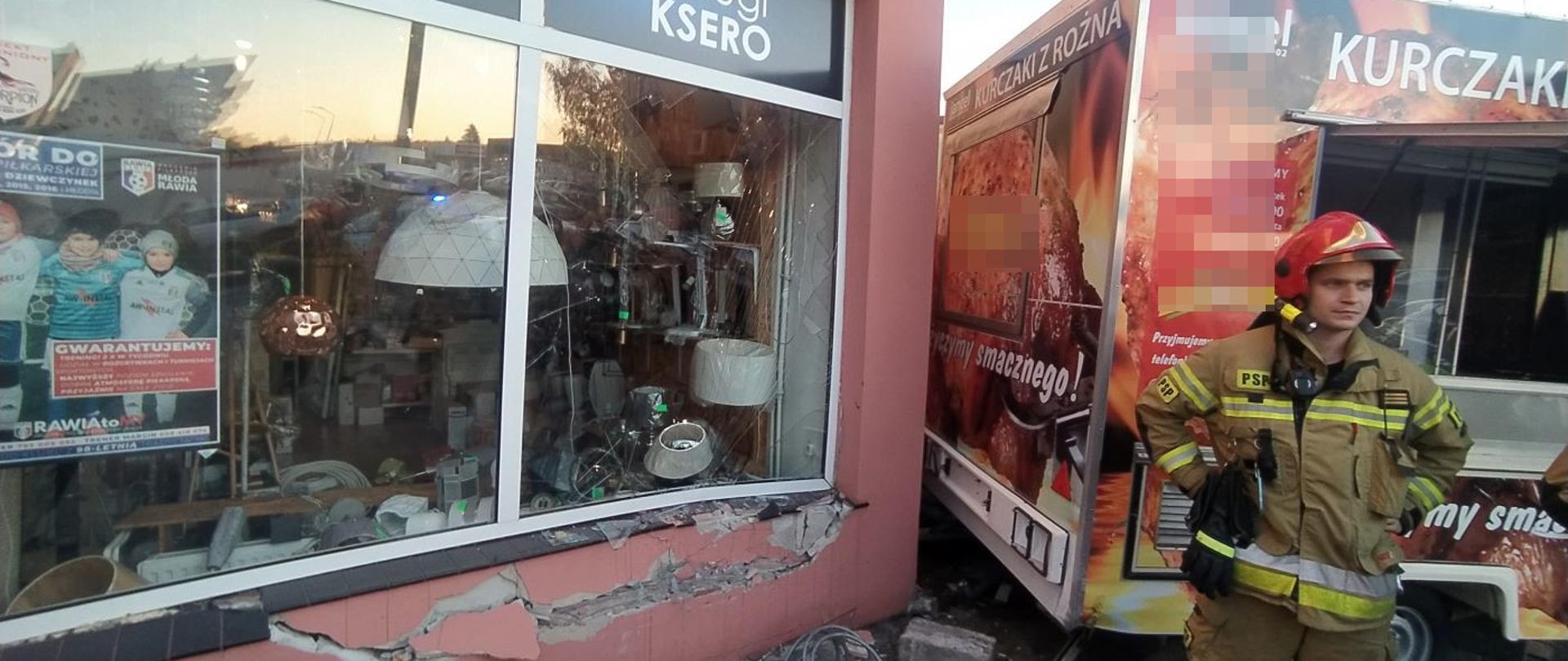 Kolizja z przyczepą gastronomiczną. Parking przed kompleksem handlowo-usługowym na ul. Westerplatte w Rawiczu. Samochód osobowy wjechał na chodnik uszkadzając ścianę i witrynę sklepu oraz przepchnął przyczepę gastronomiczną. Widoczne są pęknięcia muru oraz stłuczenia witryny sklepowej. Obok sklepu stoi przyczepa gastronomiczna. Przed nią strażak.