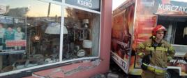 Kolizja z przyczepą gastronomiczną. Parking przed kompleksem handlowo-usługowym na ul. Westerplatte w Rawiczu. Samochód osobowy wjechał na chodnik uszkadzając ścianę i witrynę sklepu oraz przepchnął przyczepę gastronomiczną. Widoczne są pęknięcia muru oraz stłuczenia witryny sklepowej. Obok sklepu stoi przyczepa gastronomiczna. Przed nią strażak.