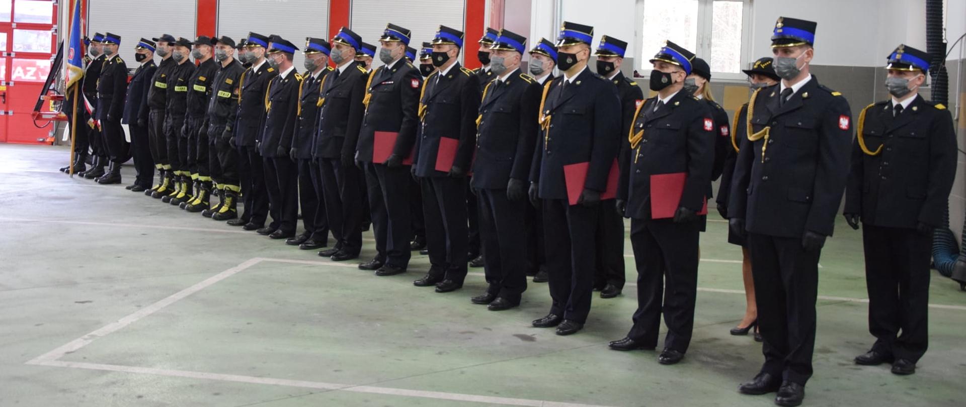 Strażacy w umundurowaniu galowym oraz ubraniach koszarowych stoją w dwuszeregu. Czworo funkcjonariuszy trzyma w dłoniach bordowe teczki. Po lewej stronie stoi poczet sztandarowy.