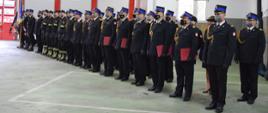 Strażacy w umundurowaniu galowym oraz ubraniach koszarowych stoją w dwuszeregu. Czworo funkcjonariuszy trzyma w dłoniach bordowe teczki. Po lewej stronie stoi poczet sztandarowy.