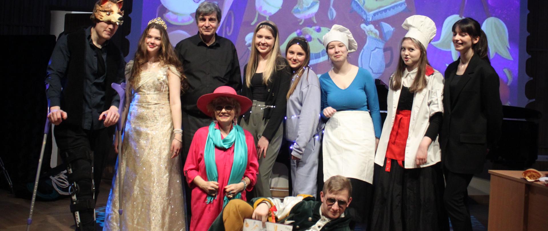 Grafika - zdjęcie. Grupa aktorów po spektaklu na estradzie pozuje do wspólnego zdjęcia. W tle fragment scenografii wyświetlony na ekranie.