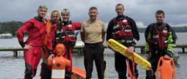 Na zdjęciu widać sześciu strażaków stojących na drewnianym pomoście nad jeziorem, po zakończonym szkoleniu. Czterech z nich ubranych jest w kombinezony do ratownictwa wodnego, w tym dwóch w czerwone skafandry z maskami i rurkami do nurkowania, a dwóch w czarno-czerwone kombinezony z kamizelkami ratunkowymi z napisem „STRAŻ”. Jeden mężczyzna w środku nosi beżową koszulkę i czarne spodnie z odblaskowymi elementami, instruktor. Drugi od lewej trzyma w rękach pomarańczowego manekina ratowniczego, kolejny z prawej trzyma żółtą deskę ratunkową z napisem „RESCUE”, a ostatni z prawej – pomarańczową bojkę ratunkową. W tle widać jezioro, pomosty oraz zachmurzone niebo, co sugeruje, że zdjęcie wykonano podczas szkolenia i egzaminu z ratownictwa wodnego.