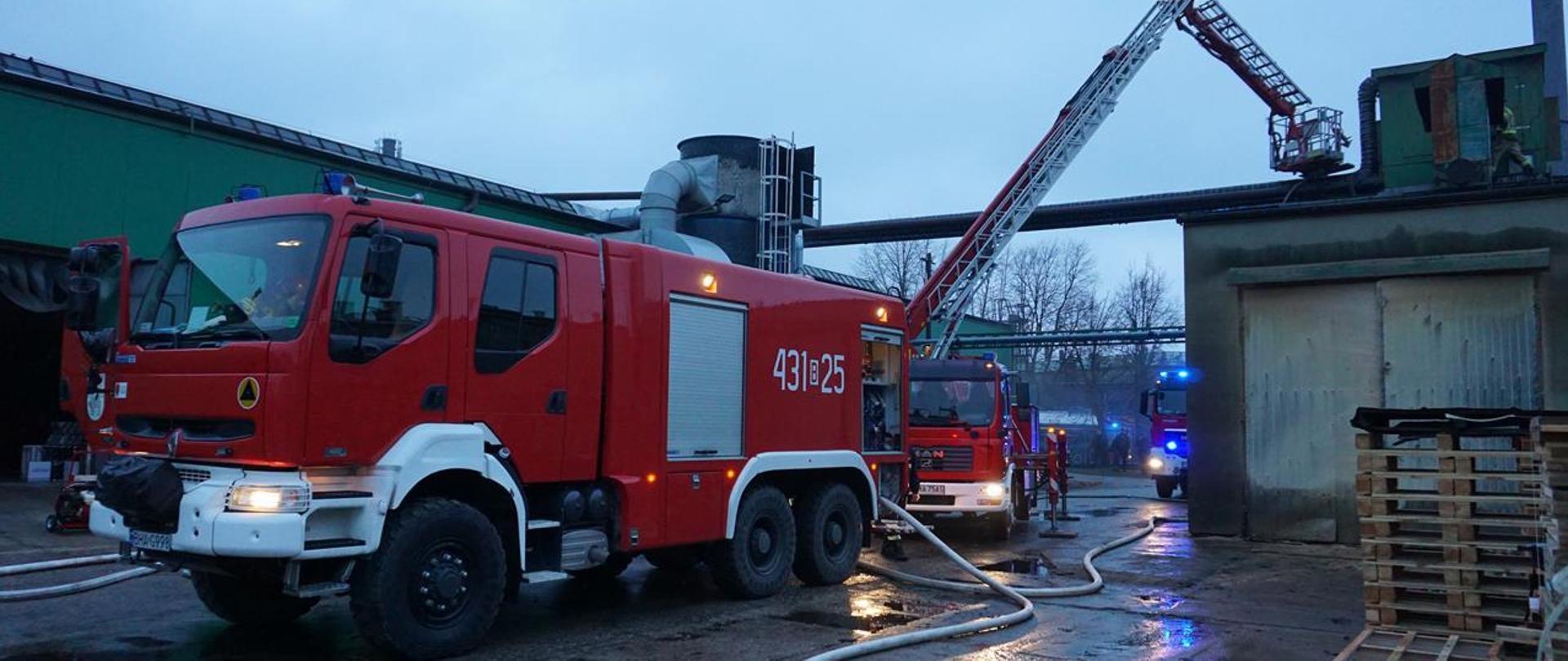 Na pierwszym planie stoi czerwony samochód pożarniczy z podpiętymi wężami pożarniczymi, na planie drugim widać rozłożony, pożarniczy podnośnik hydrauliczny.