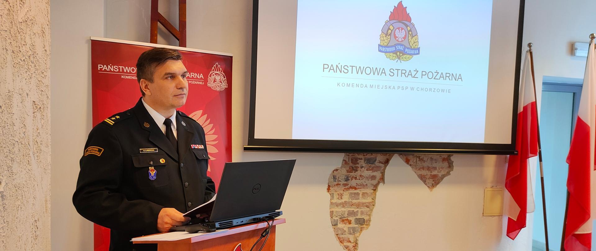Zdjęcie przedstawia Komendanta Miejskiego PSP w Chorzowie - bryg. Arkadiusza Labochę, stojącego przy mównicy na tle ekranu projekcyjnego