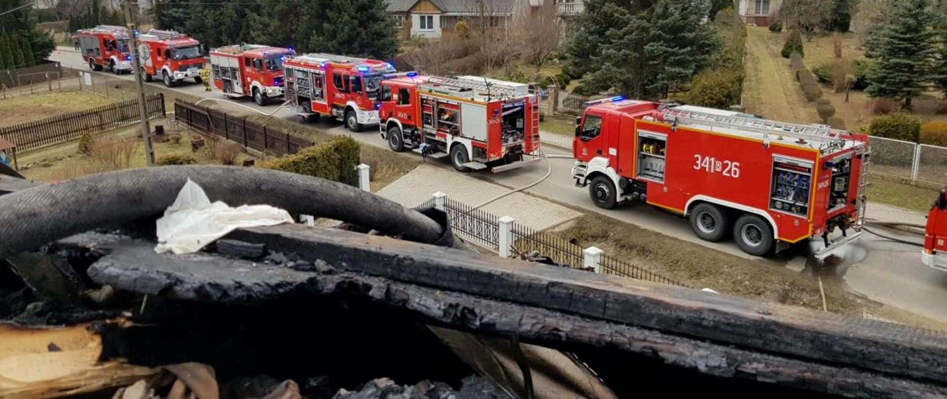 Zdjęcie wykonano z poddasza spalonego budynku mieszkalnego w kierunku zaparkowanych na ulicy ciężkich samochodów gaśniczych. W oddali widać 7 samochodów Państwowej Straży Pożarnej oraz ochotniczych straży pożarnych. W rogu zdjęcia uwidocznione są spalone krokwie stanowiące konstrukcję dachu