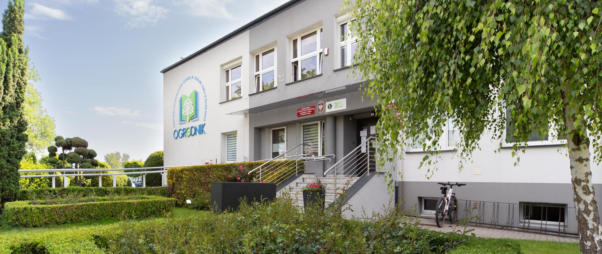 Dwukondygnacyjny jasnoszary budynek z logo „OGRODNIK” na elewacji. Przed wejściem znajduje się zadbany ogród z geometrycznymi żywopłotami, krzewami róż i drzewami ozdobnymi. Po lewej stronie stoi rower, a na prawo rośnie brzoza. Widok w słoneczny dzień.
