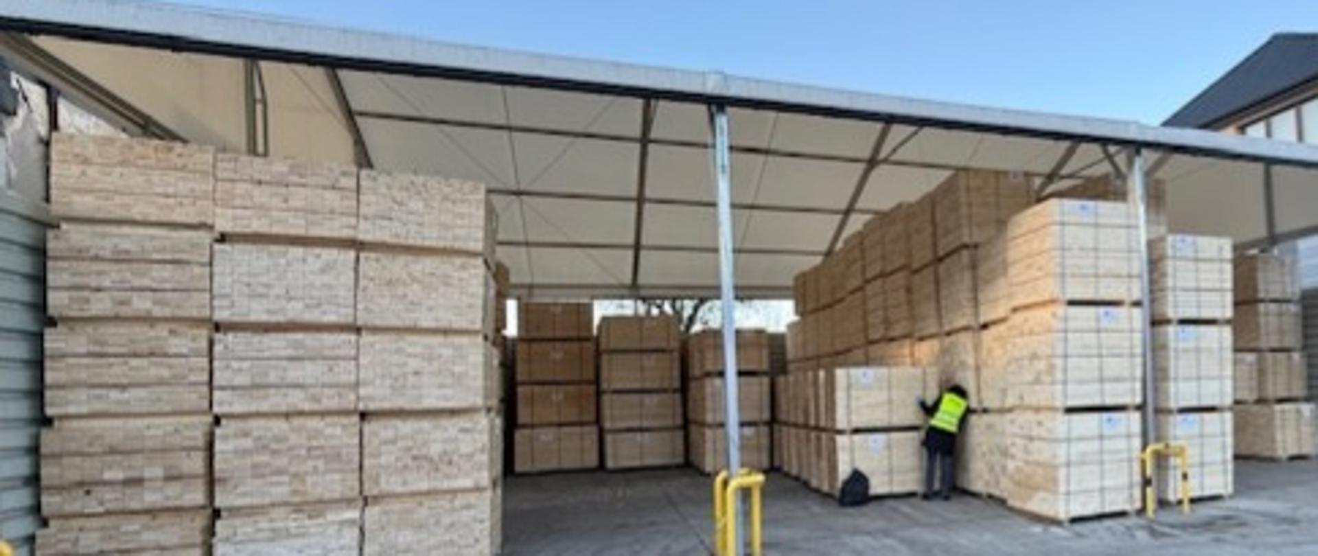 Wiata, pod którą umieszczone są elementy skrzyniopalet spakowane w postaci sześciennych pakietów. Pomiędzy pakietami widoczny inspektor PIORiN w trakcie lustracji fitosanitarnej.