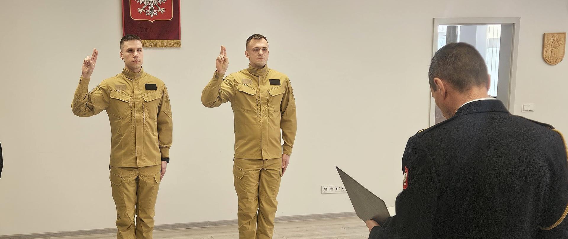 Na zdjęciu widzimy młodych mężczyzn w ubraniu koszarowym. Stoją w postawie oficjalnej, z uniesioną prawą ręką i złożonymi trzema palcami – jest to gest ślubowania, charakterystyczny dla ceremonii składania przysięgi.
W tle widoczne jest godło Polski – biały orzeł w koronie na czerwonym tle, co dodatkowo podkreśla rangę wydarzenia i jego oficjalny charakter.
