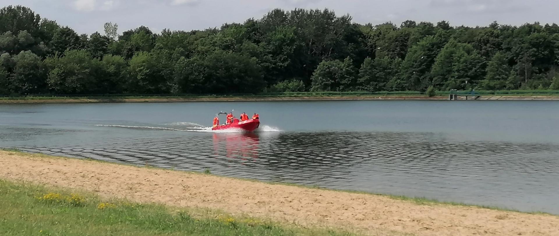 Na 1 planie trawa i piaszczysta plaża, w tle woda i strażacy wraz z Policjantem płynący łodzią. W tle drzewa. 