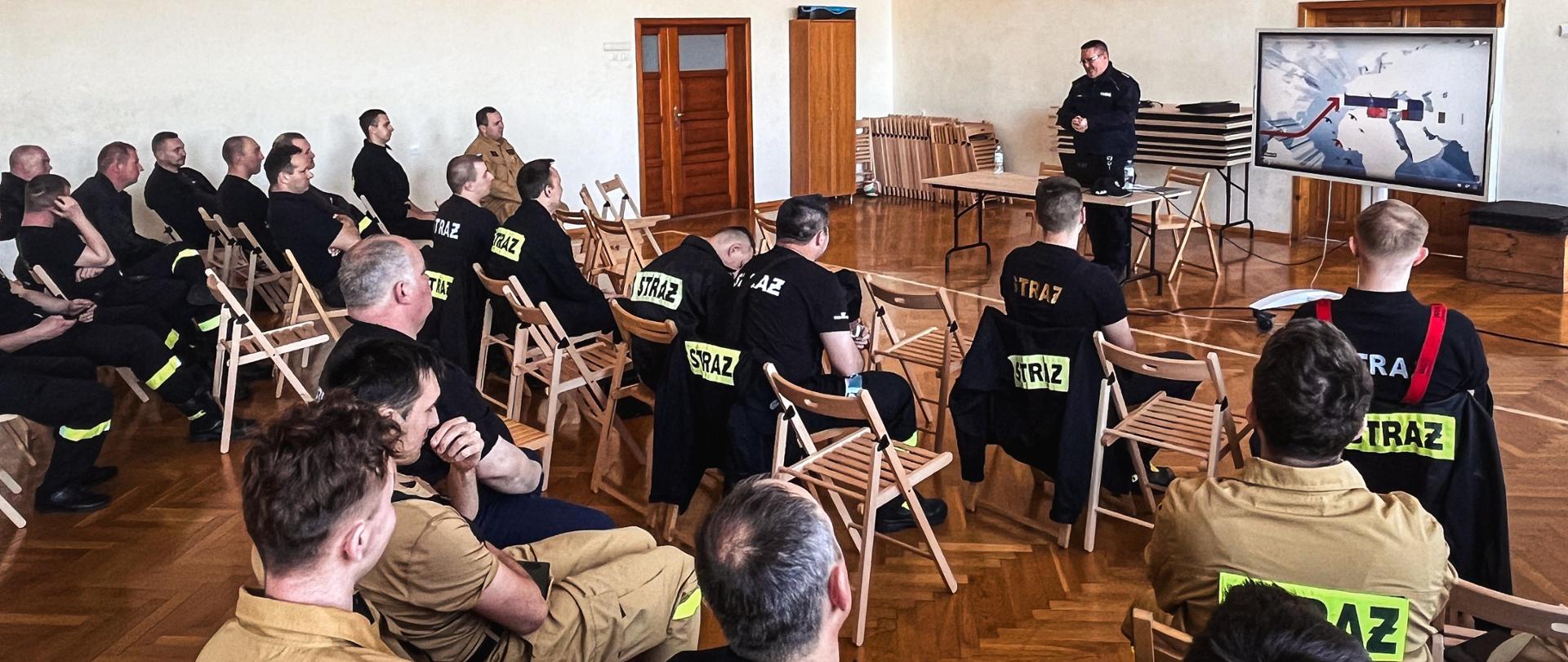 Słuchacze siedzą w sali gimnastycznej podczas prelekcji funkcjonariusza Policji. Obok prelegenta znajduje się ekran z prezentacją.