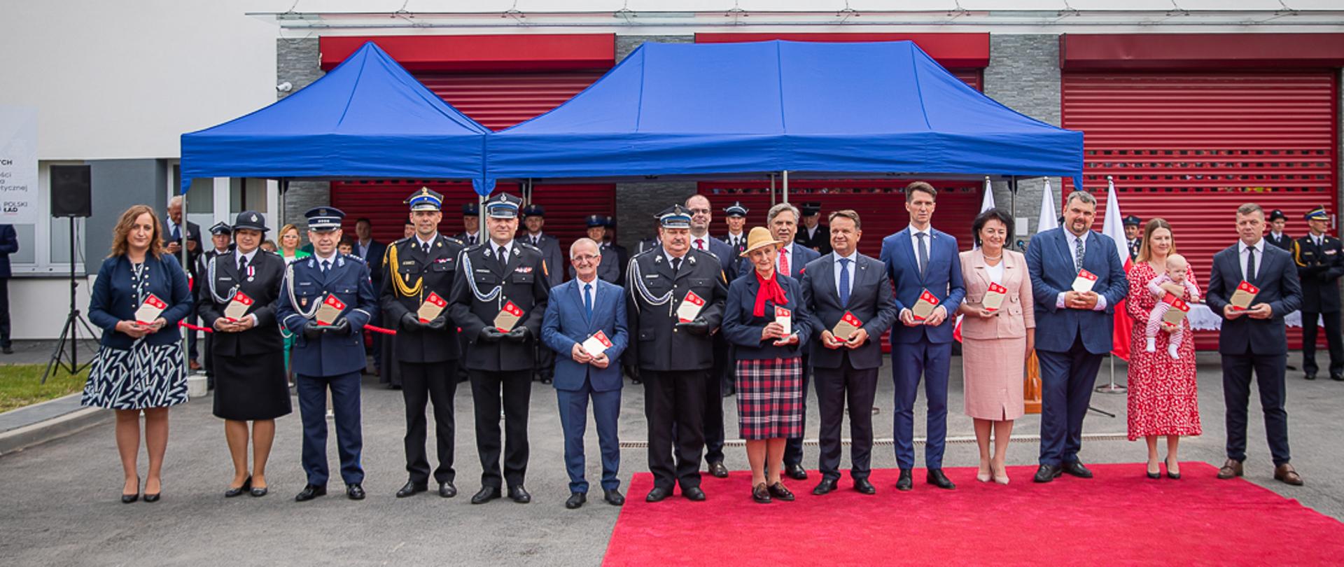 Na zdjęciu przed niebieskimi namiotem zaproszeni i wyróżnieni goście strażacy z PSP i OSP oraz osoby cywilne uczestniczące w uroczystości poświęcenie i przekazania samochodów pożarniczych dla druhów z OSP. 