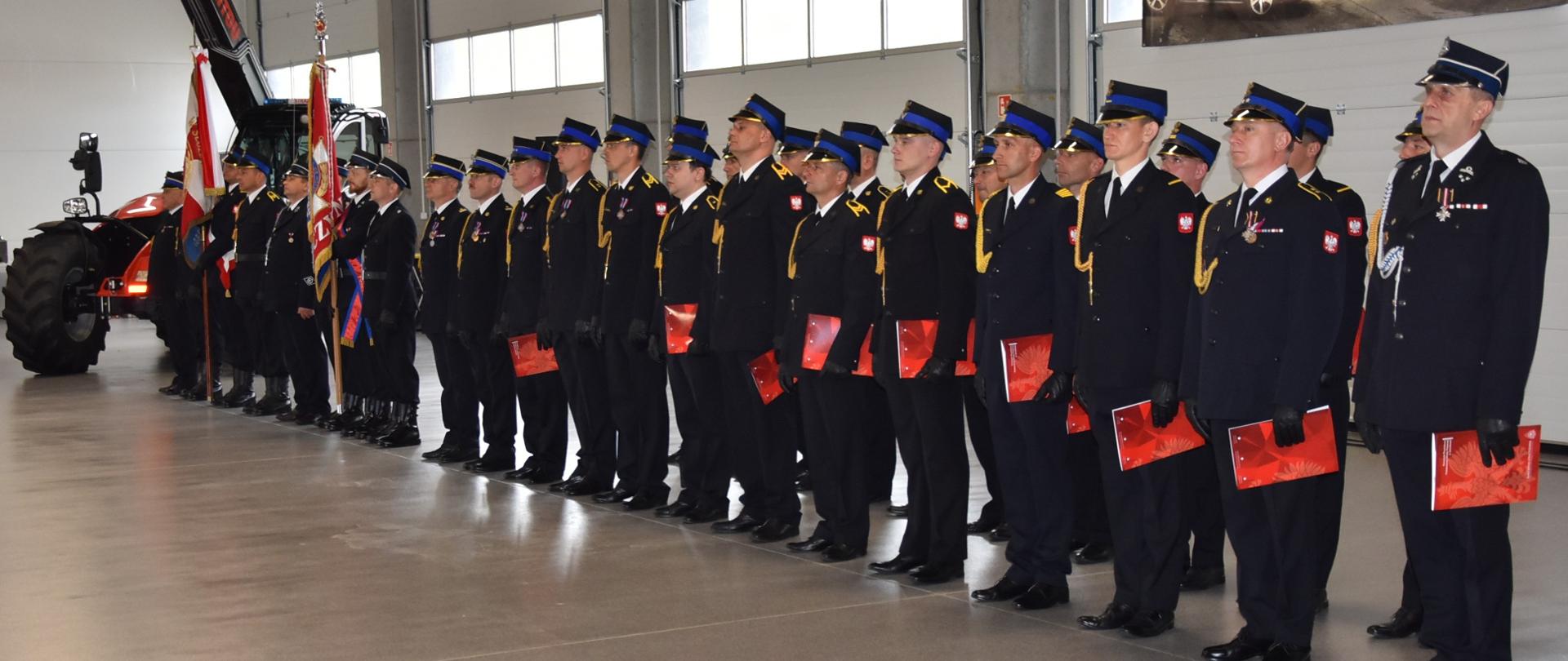Na zdjęciu znajduje się grupa osób stojących w hali jednostki straży pożarnej. Uczestnicy trzymają wyróżnienia. W tle widoczni są strażacy w mundurach galowych oraz baner Komendy Powiatowej PSP w Jarosławiu.