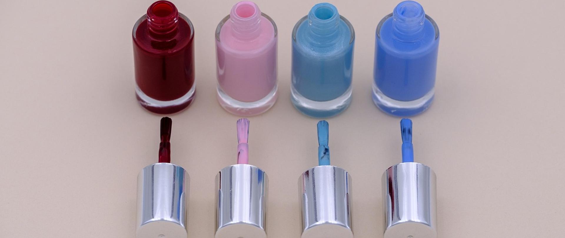 na zdjęciu cztery lakiery do paznokci ustawione w rzędzie na beżowym tle, wszystkie są odkręcone a zatyczki z pędzelkiem leżą przed nimi, pierwszy lakier od lewej jest w kolorze bordowym, drugi różowy, trzeci jasnoniebieski i czwarty niebieski