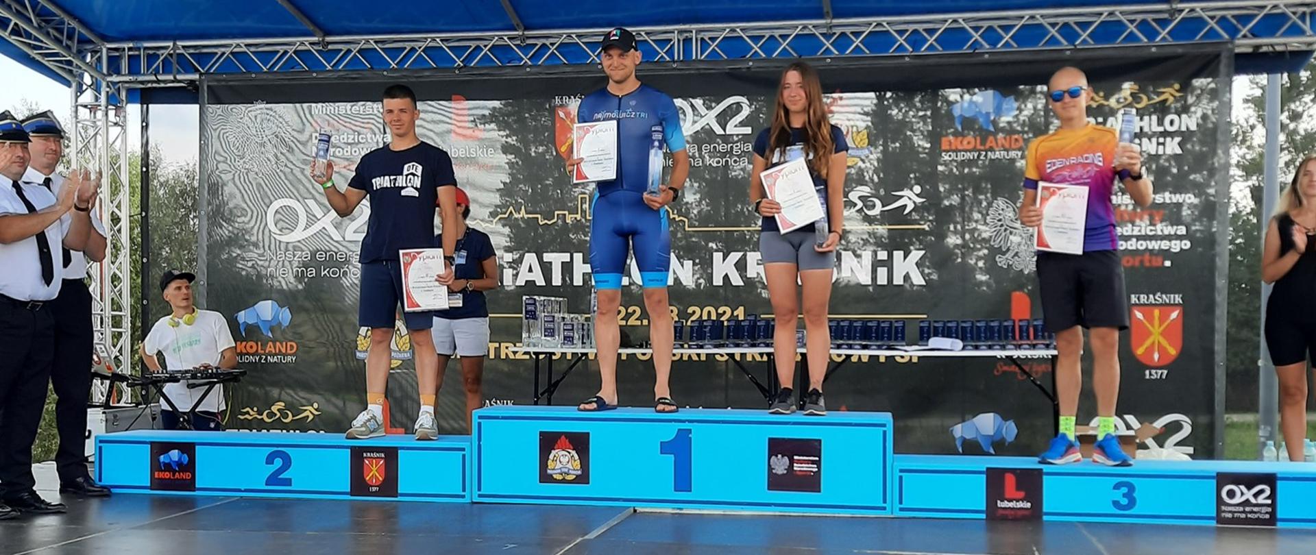 Zdjęcie wykonano porą dzienną. Przedstawia podium a na nim pierwsze trzy miejsca zawodów II Mistrzostw Polski Strażaków w Triathlonie. Zawodnicy w strojach sportowych po lewej stronie dwóch strażaków w Mundurach galowych. 