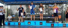Zdjęcie wykonano porą dzienną. Przedstawia podium a na nim pierwsze trzy miejsca zawodów II Mistrzostw Polski Strażaków w Triathlonie. Zawodnicy w strojach sportowych po lewej stronie dwóch strażaków w Mundurach galowych. 