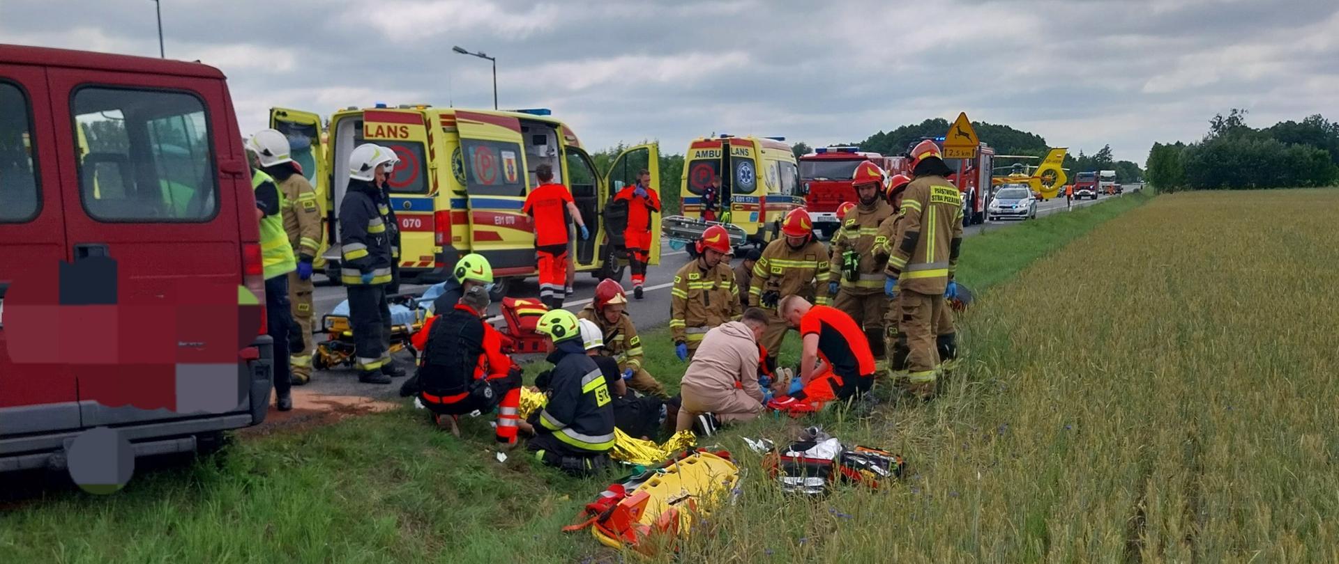 Zdjęcie przedstawia wypadek drogowy. Na pierwszym planie widzimy strażaków, ratowników medycznych oraz osoby udzielające pomocy poszkodowanym leżącym na trawie. Ratownicy używają sprzętu medycznego, obok nich na trawie leżą torby ratownicze. W tle widzimy ambulanse, wozy strażackie oraz czerwony bus, który brał udział w wypadku. 